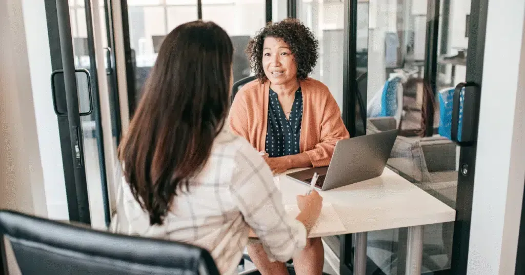 Two people in a glass-walled office discussing work across a conference table with a laptop.