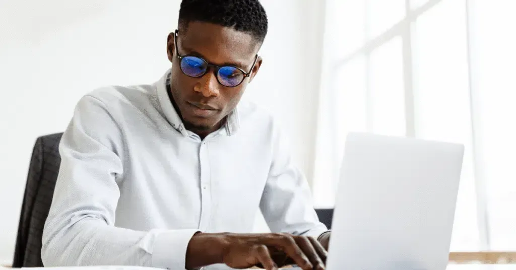 Man in a white shirt and blue sunglasses typing on a laptop at a bright desk