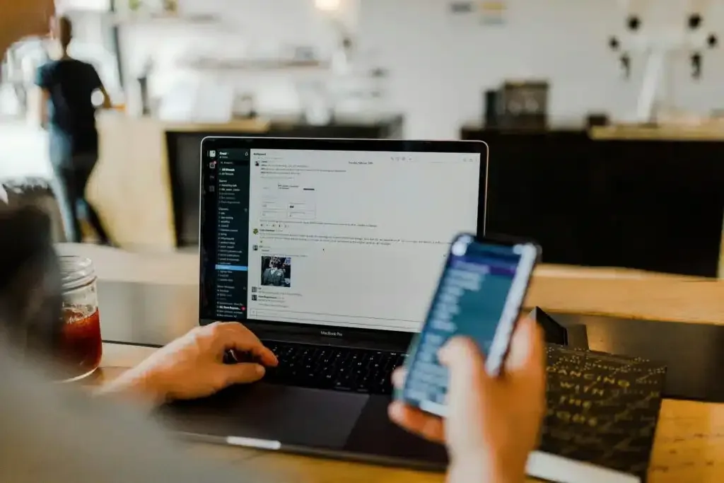 Person using a laptop and smartphone at a café table, with a blurred background.