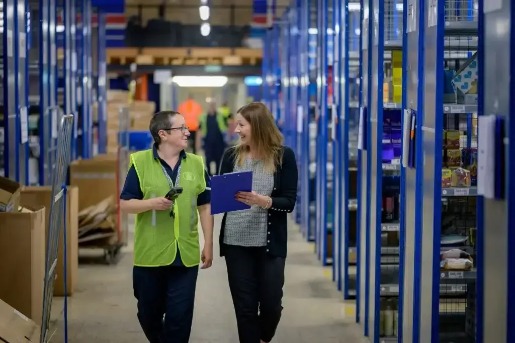 Two coworkers walk through a warehouse aisle, talking while reviewing a clipboard.