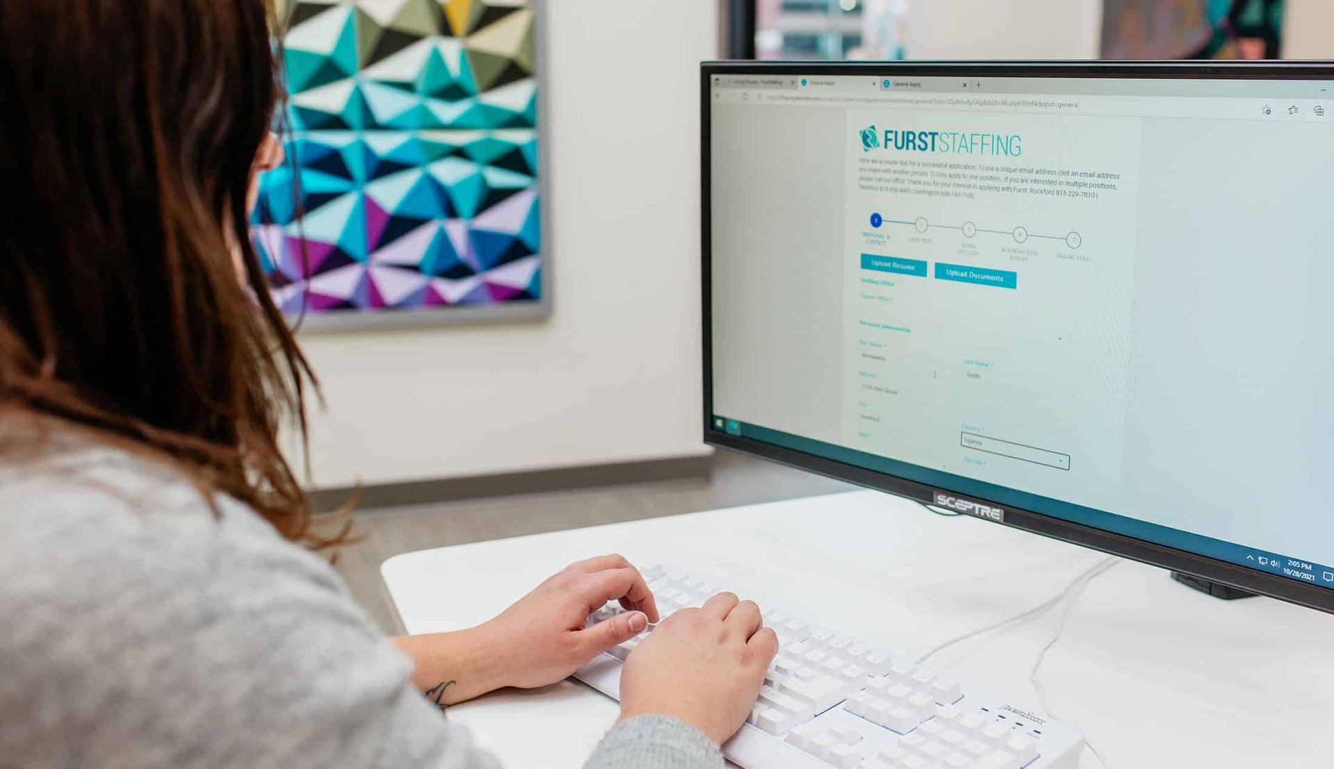 Person typing at a desktop computer in a bright office, viewing a website on the monitor.