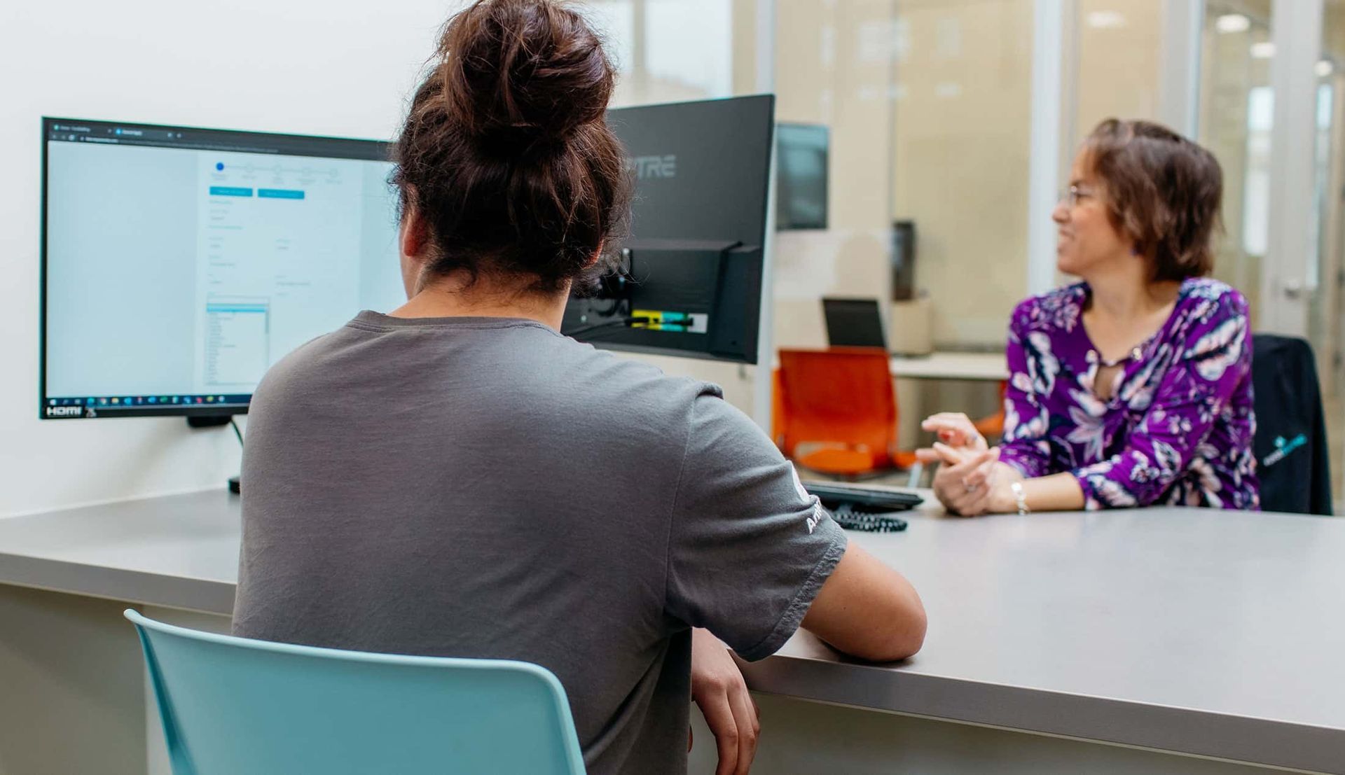 Two people talking across a desk in an office, with a computer monitor and paperwork visible.