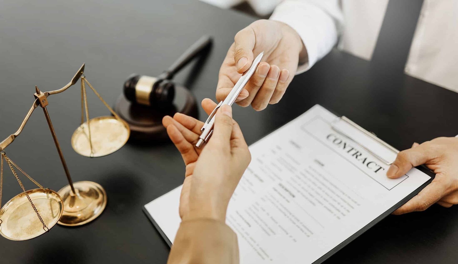 Hands exchanging a pen over a contract on a desk with a scale of justice and gavel