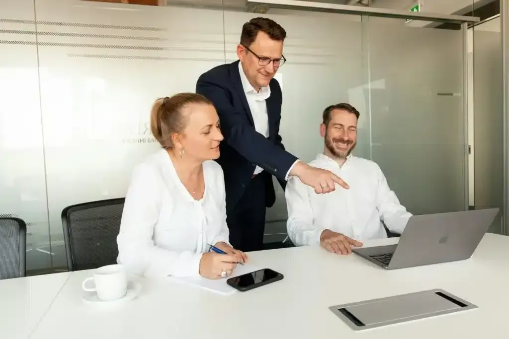 Three coworkers smiling in a modern office, gathered around a laptop during a meeting