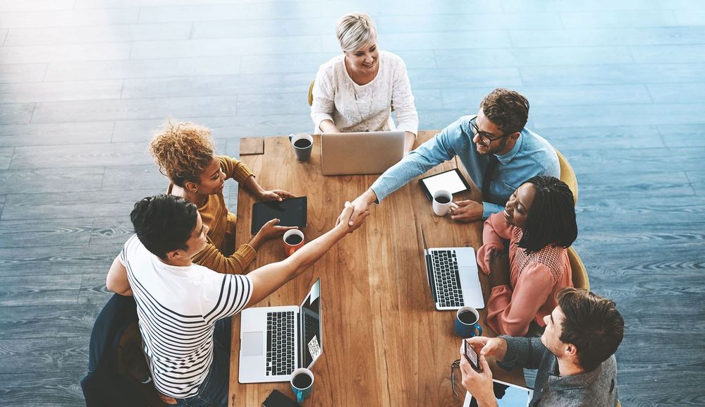 Team gathered around a wooden table in a bright office, collaborating with laptops and coffee cups.
