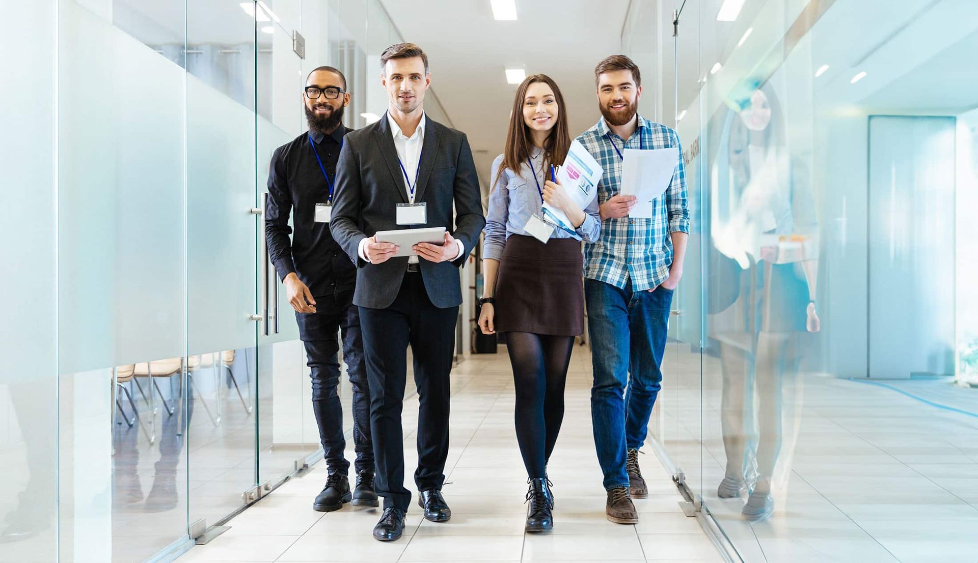 Four coworkers walking through a bright office hallway, one holding documents, with glass walls on both sides