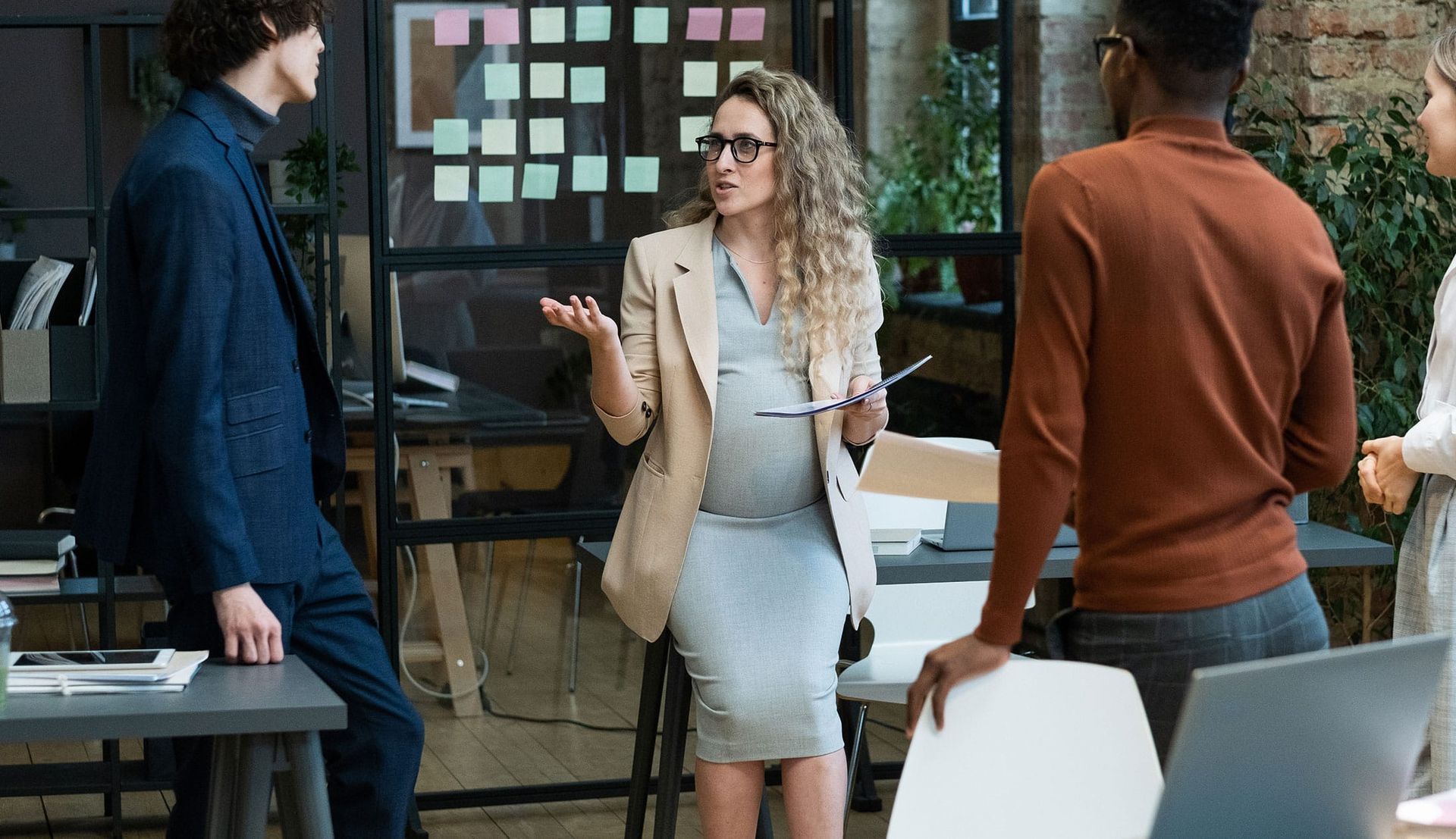Three coworkers talking in a modern office, with one woman holding papers and gesturing.