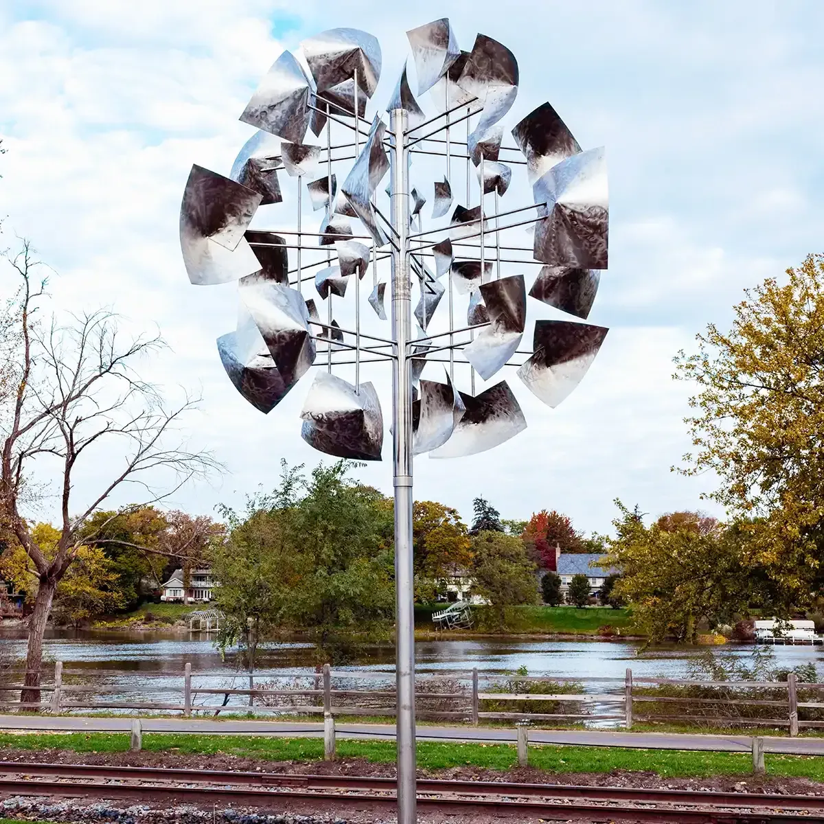 Metal wind sculpture by a riverside path with autumn trees and a cloudy sky