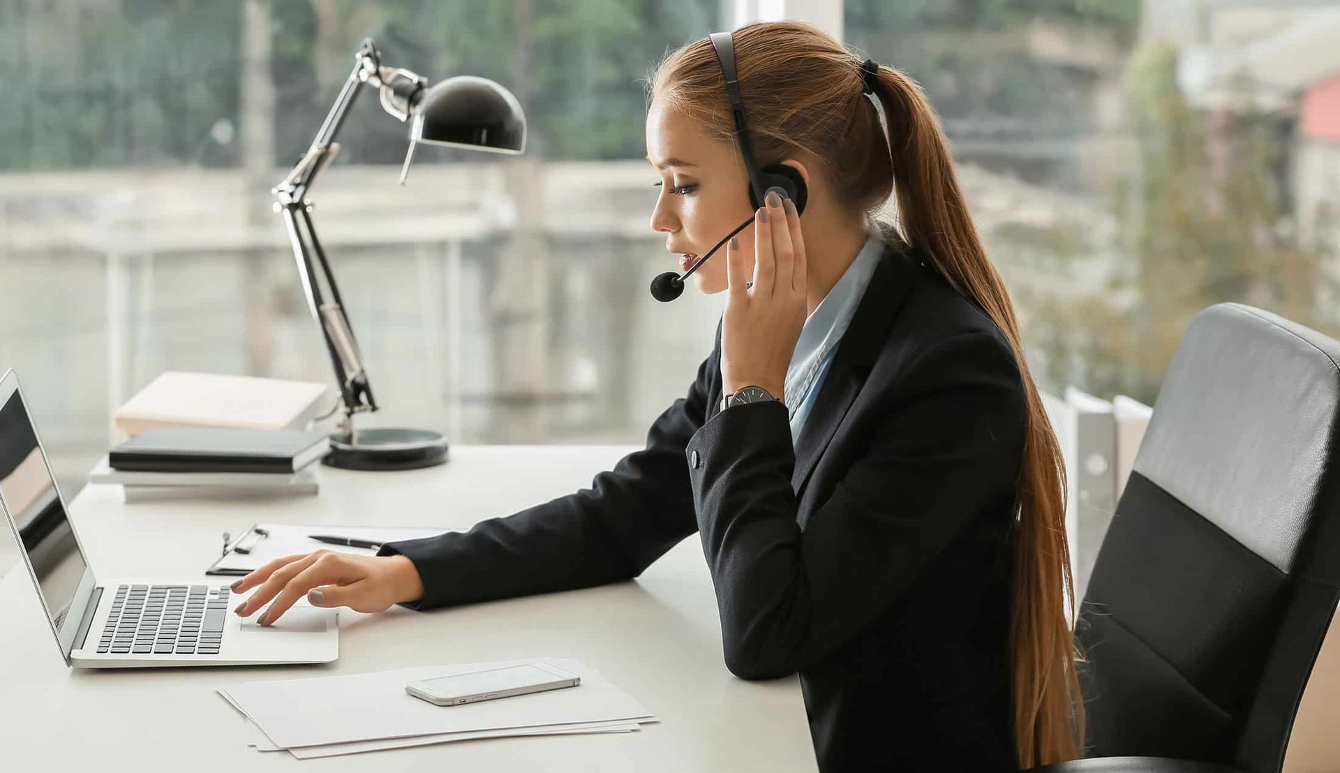 Woman on a phone call working at a desk with a laptop and lamp in a bright office