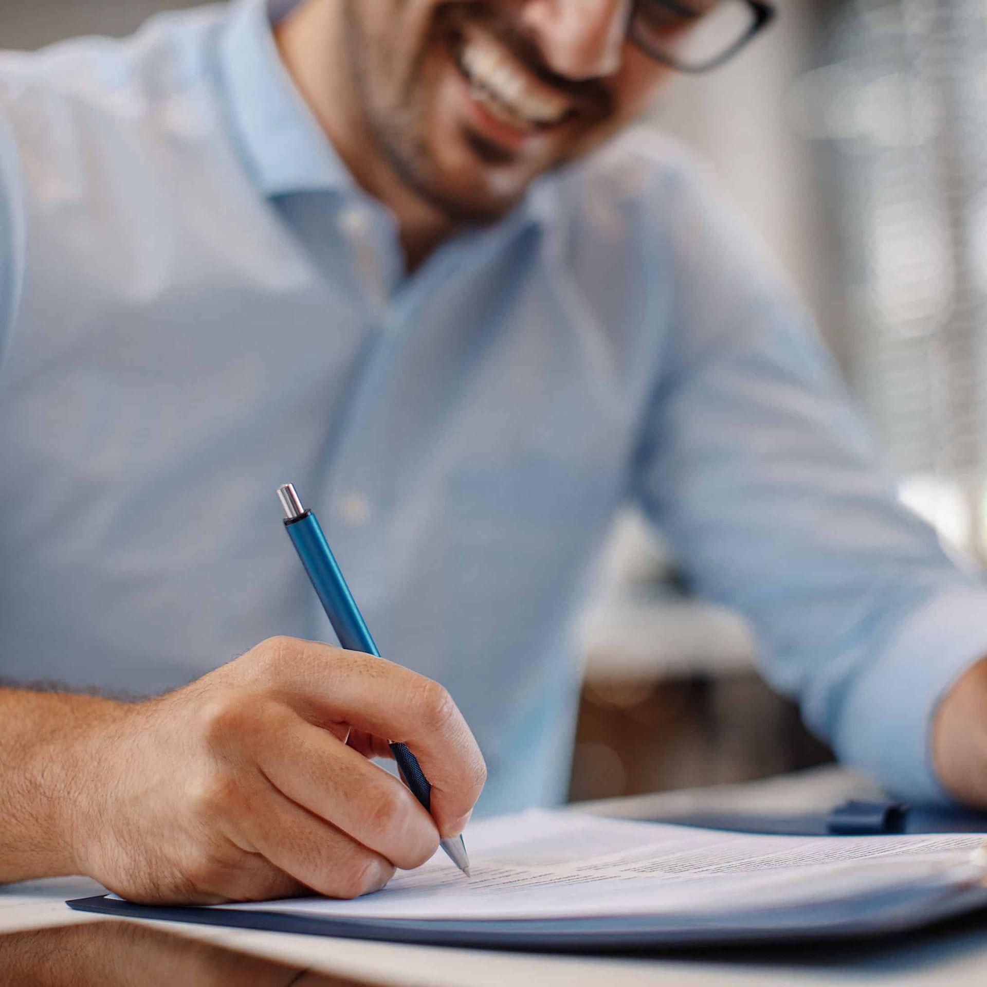 Person writing in a notebook with a blue pen at a desk, smiling in a bright office setting