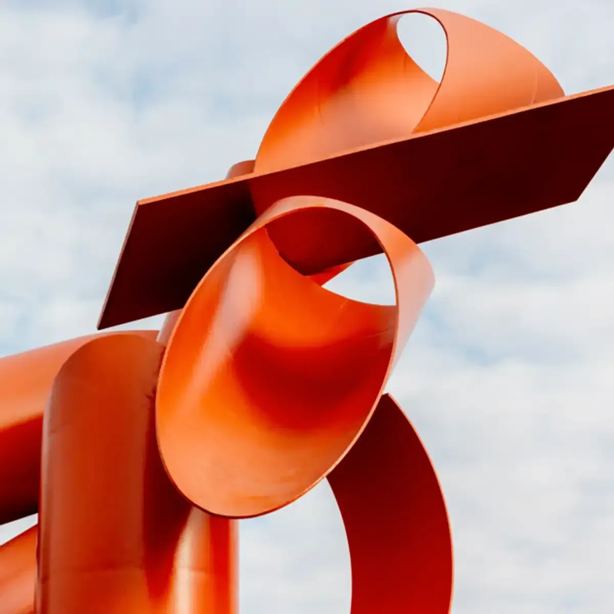 Abstract orange-red metal sculpture against a cloudy sky
