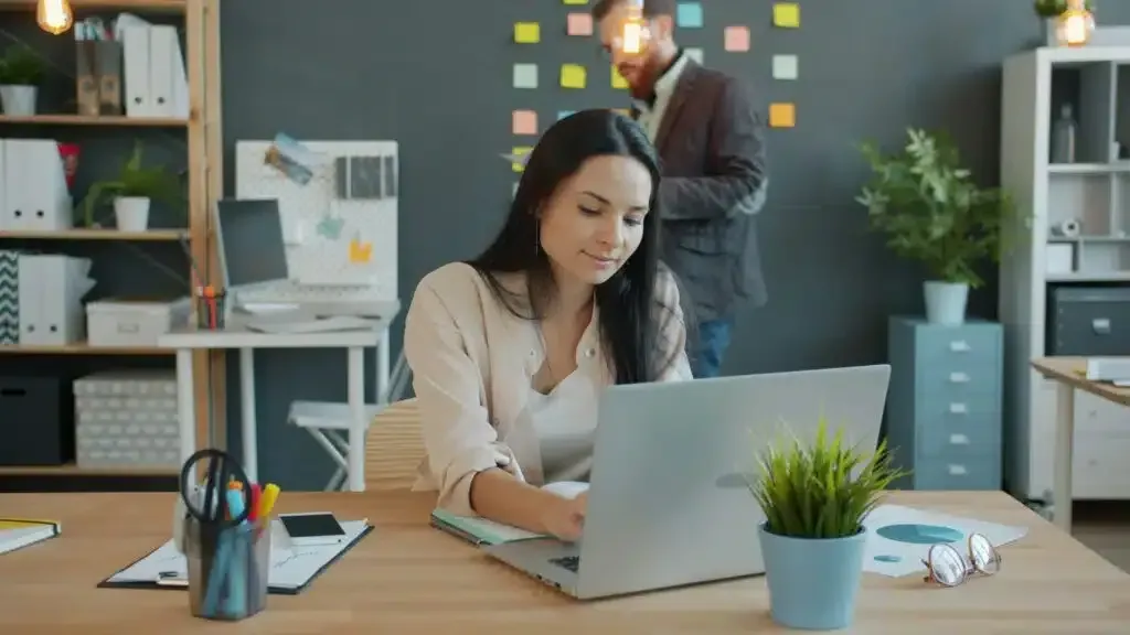 Person working on a laptop at a desk in a modern office, with a coworker in the background.