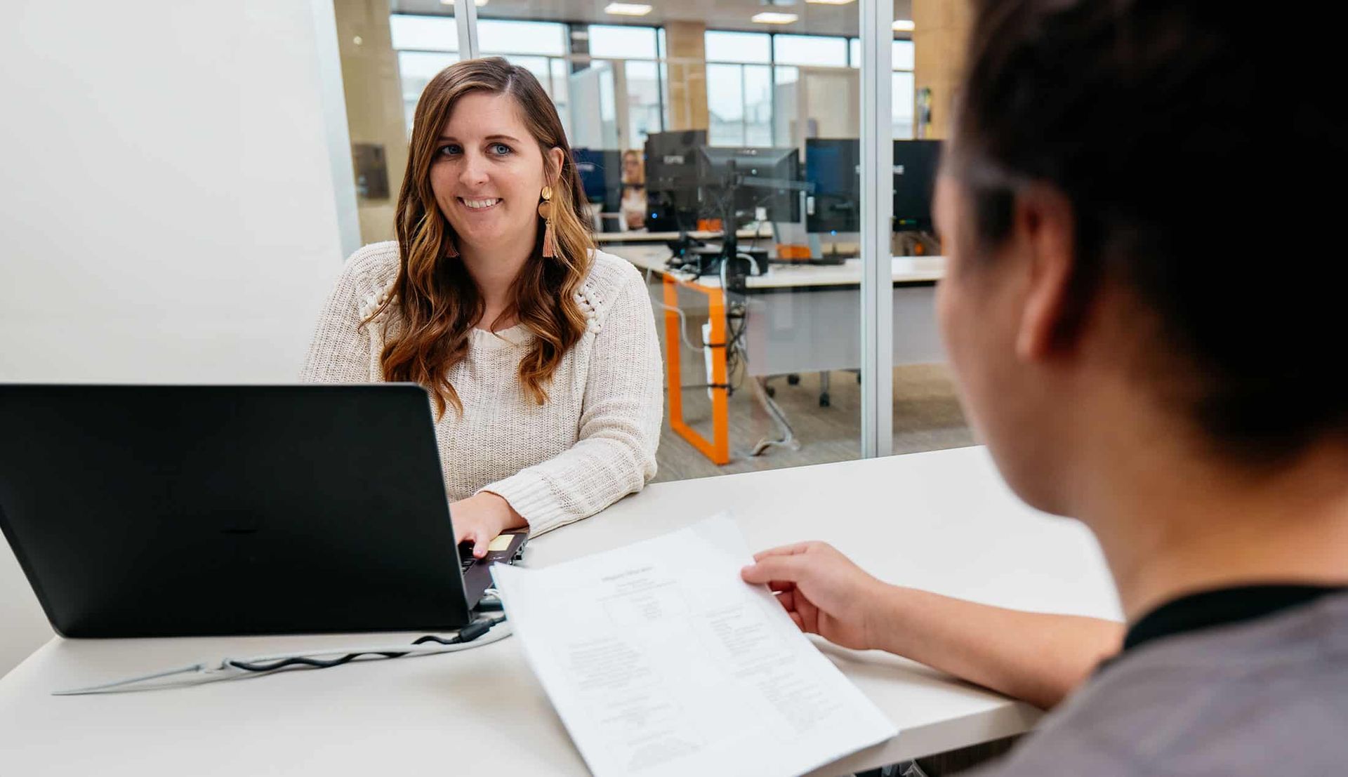 Two people in a bright office meeting across a laptop, with papers on the desk.