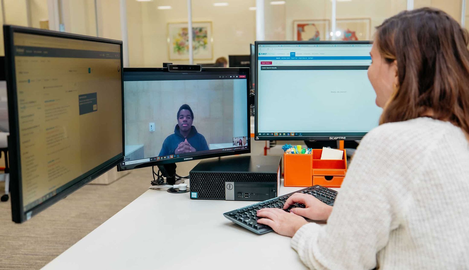 Person typing at a desk with three monitors, including a video call and a chat window in an office