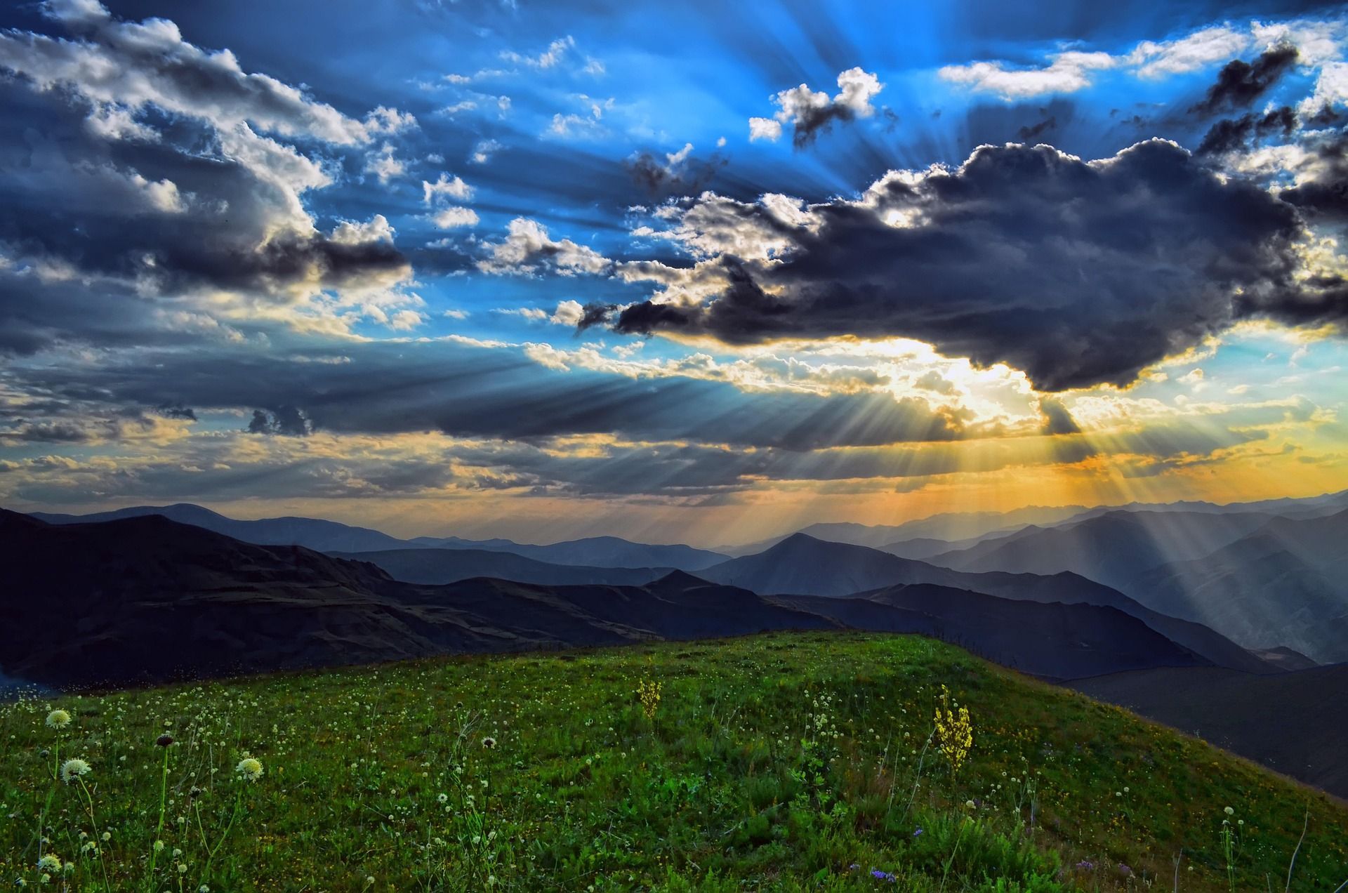 Sunbeams break through dramatic clouds over a vibrant green mountain meadow during sunset, illuminating the hilly landscape.