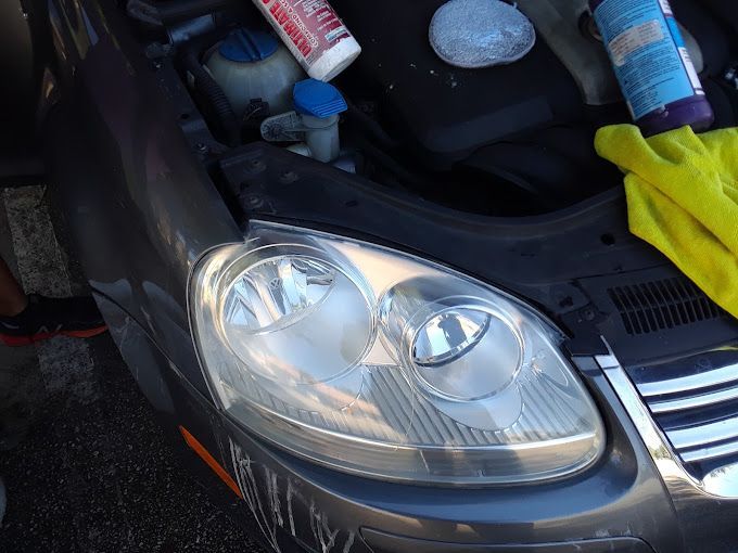 Car headlight being cleaned; gray car, cleaning supplies, focus on the headlight.