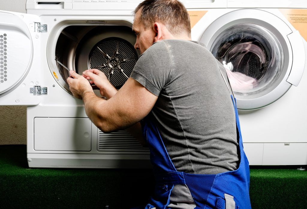 Man in blue overalls repairing a white dryer with tools.