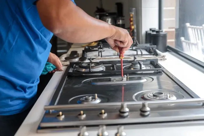 Person repairs a gas stovetop with a screwdriver in a kitchen near a window.