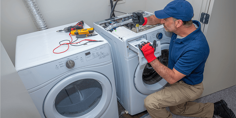 A repairman in a blue shirt and baseball cap fixing a washing machine in a laundry room, with tools visible.