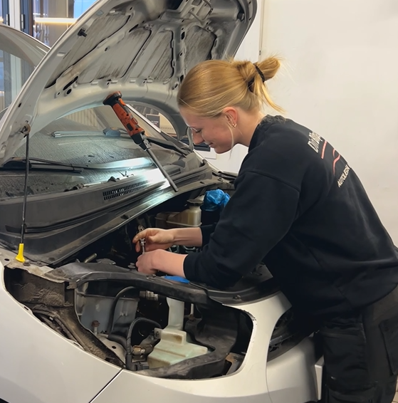 Kvinde i sort skjorte arbejder på en bilmotor med motorhjelmen oppe.