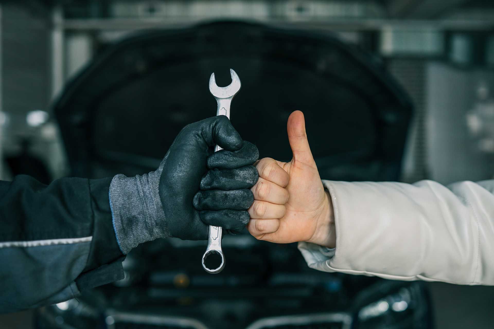 Mekaniker holder skruenøgle med behandsket hånd, kunde giver tommelfingeren op nær åben motorhjelm.