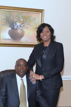 Two people posing indoors beside a framed floral painting, one seated and one standing with hands on his chair.