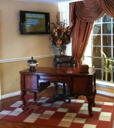 Elegant home office with wooden desk, wall-mounted TV, floral arrangement, and large curtained window