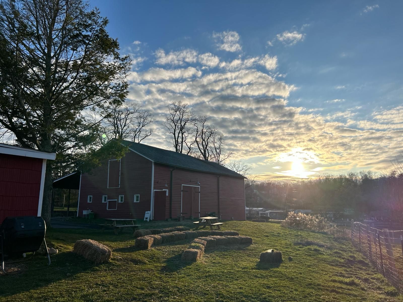 Red barn in a grassy field with hay bales, under a sky with clouds and the setting sun.