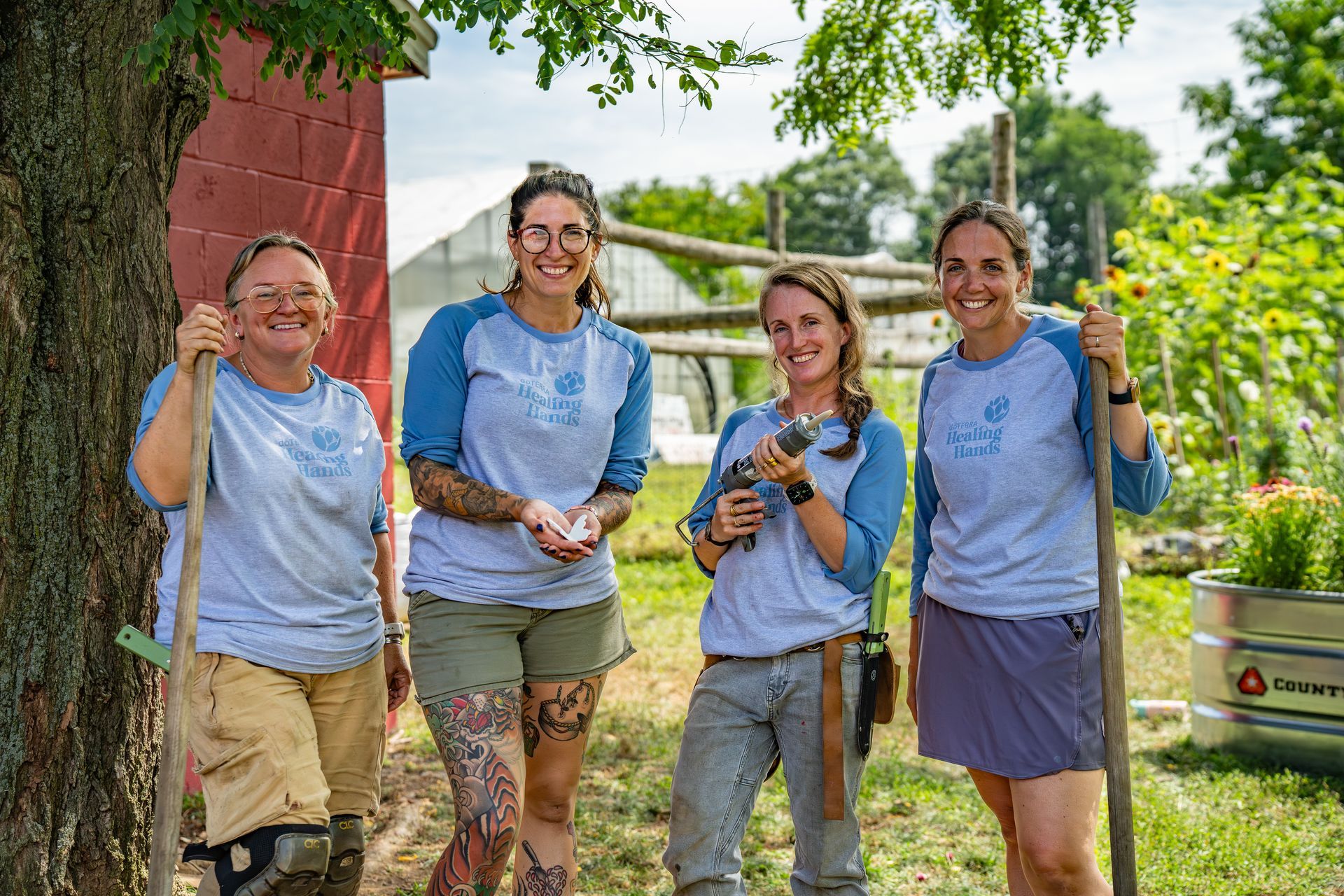 Four people in blue shirts pose outdoors with gardening tools. Greenery, a red building, and a trailer are in the background.