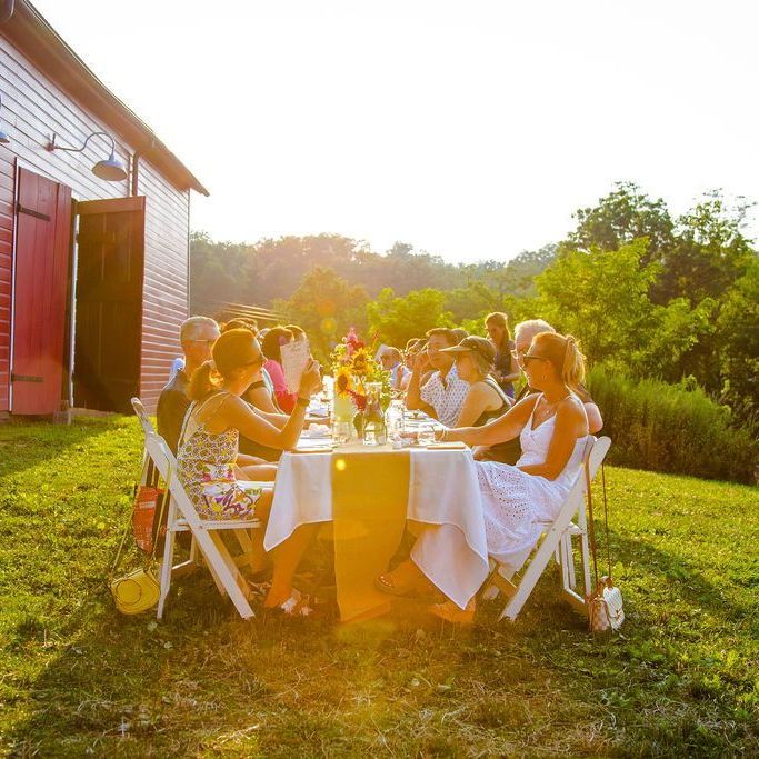 People seated at a long table outdoors near a red barn; sunny day, sunflowers, white table cloth, relaxed gathering.