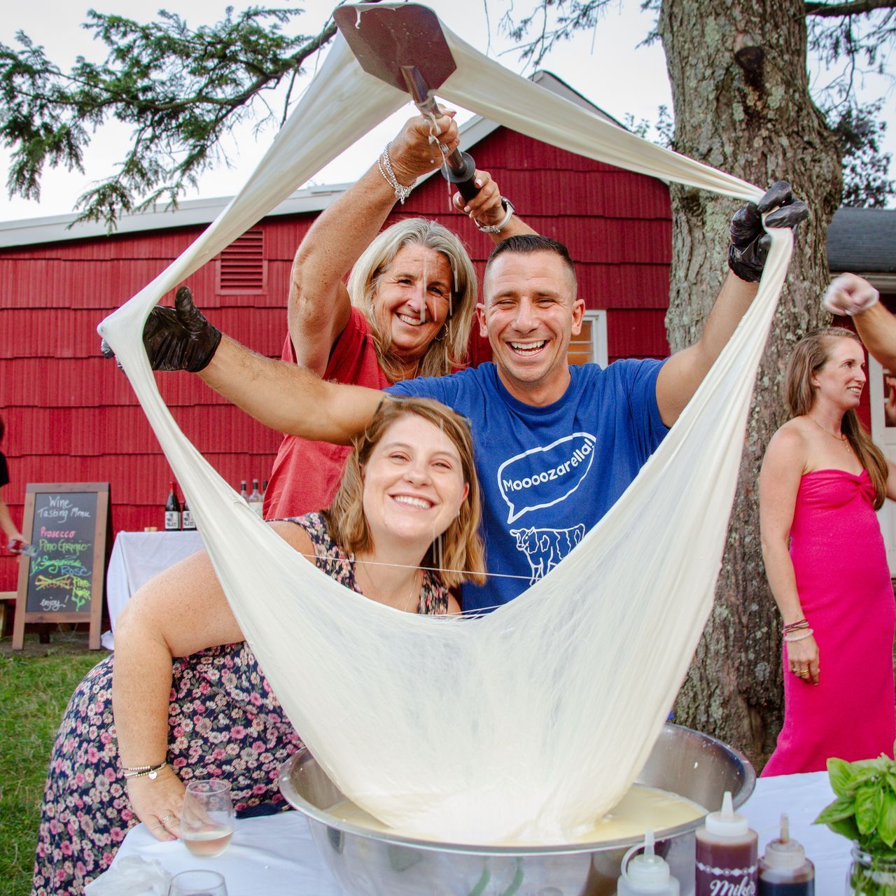 Three people stretching a large piece of cheese outdoors; a red barn is in the background.