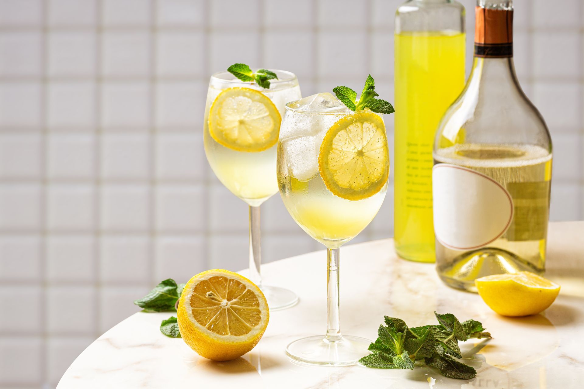 Two lemon cocktails with mint garnish, next to lemons and a wine bottle on a marble table.