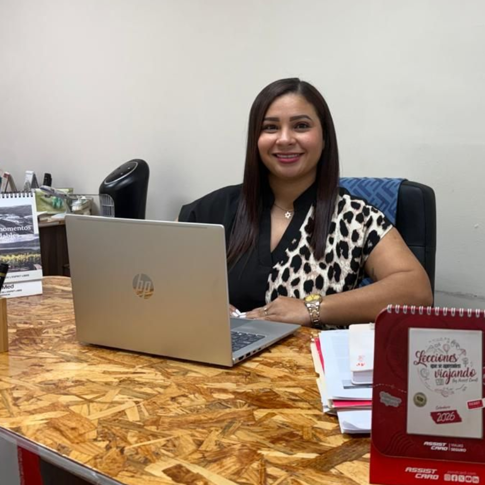 Mujer sonriendo en un escritorio con una computadora portátil, papeles y una placa roja de premio en un entorno de oficina.