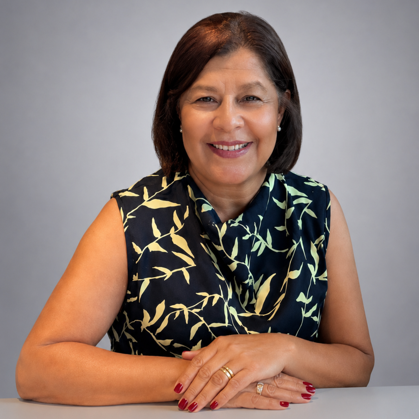 Persona sonriente sentada a una mesa, con una blusa azul marino sin mangas con estampado de hojas color crema.