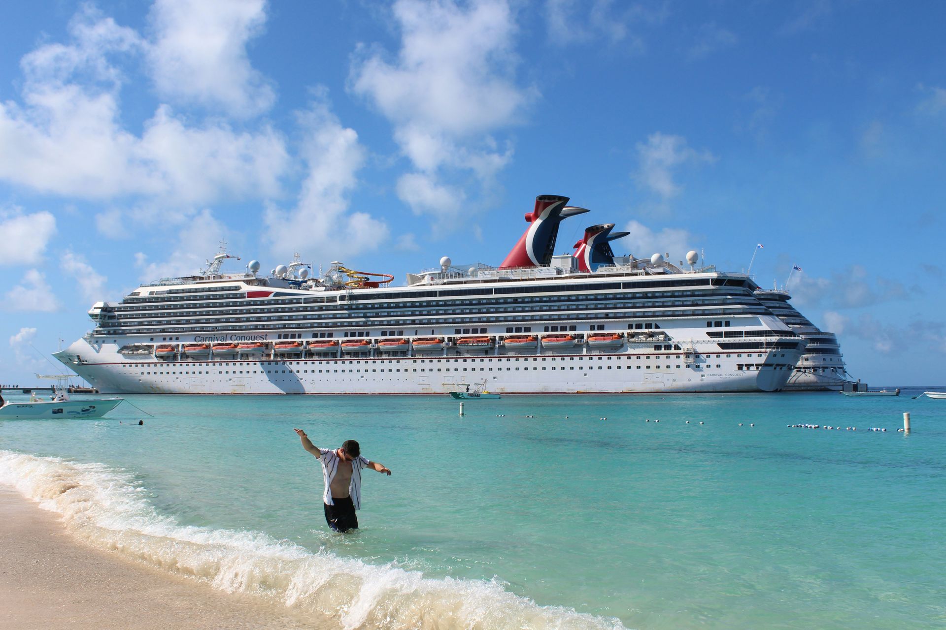 Crucero cerca de una playa tropical, con una persona de pie en aguas turquesas poco profundas bajo un cielo azul.