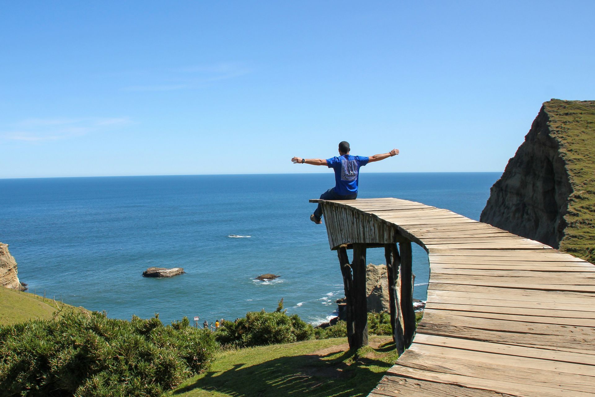 Persona de pie con los brazos extendidos en una pasarela de madera al borde de un acantilado con vistas al océano.