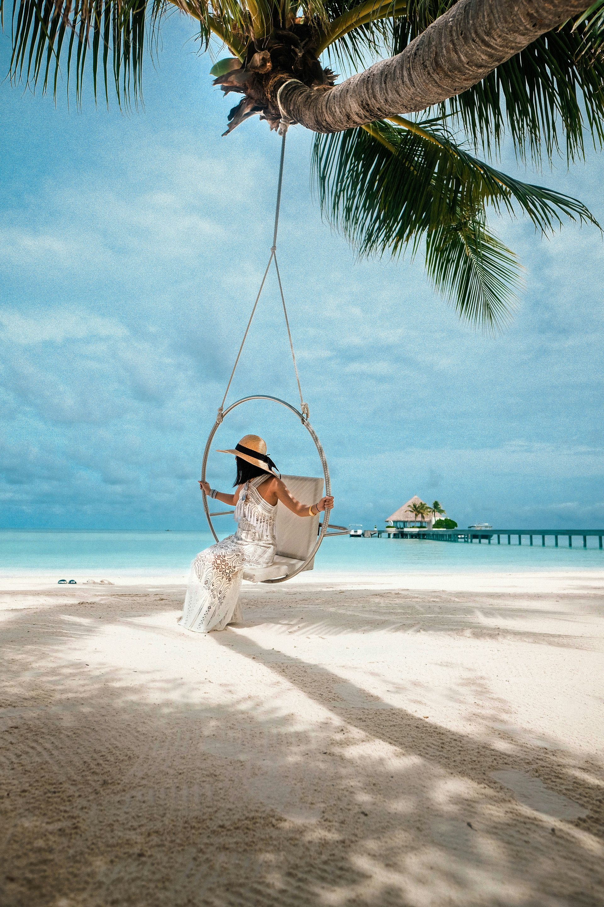 Mujer con vestido blanco sentada en un columpio colgante bajo una palmera en una playa tropical.