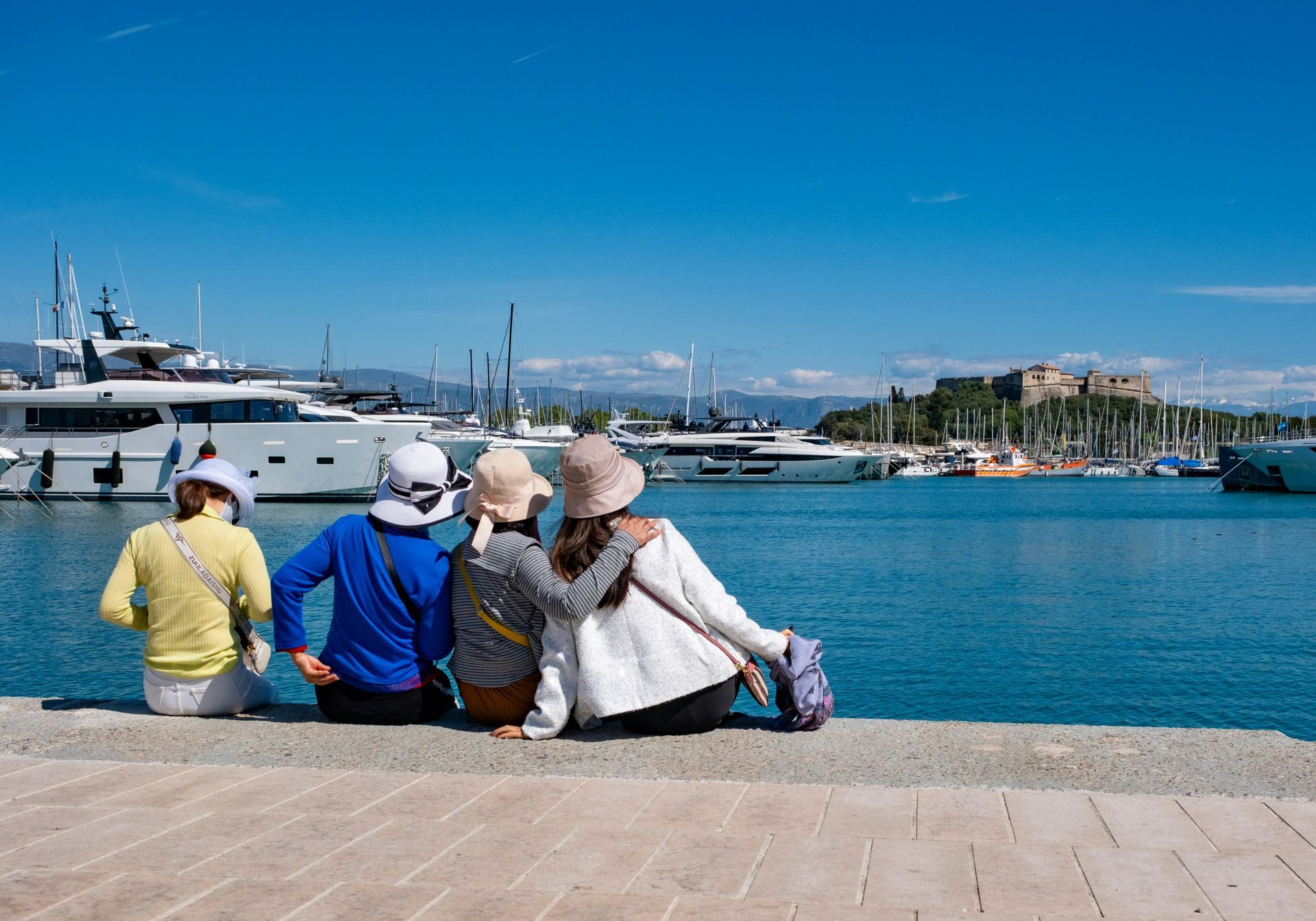 Cuatro personas están sentadas junto a un puerto deportivo, contemplando el agua azul y los yates bajo un cielo despejado.