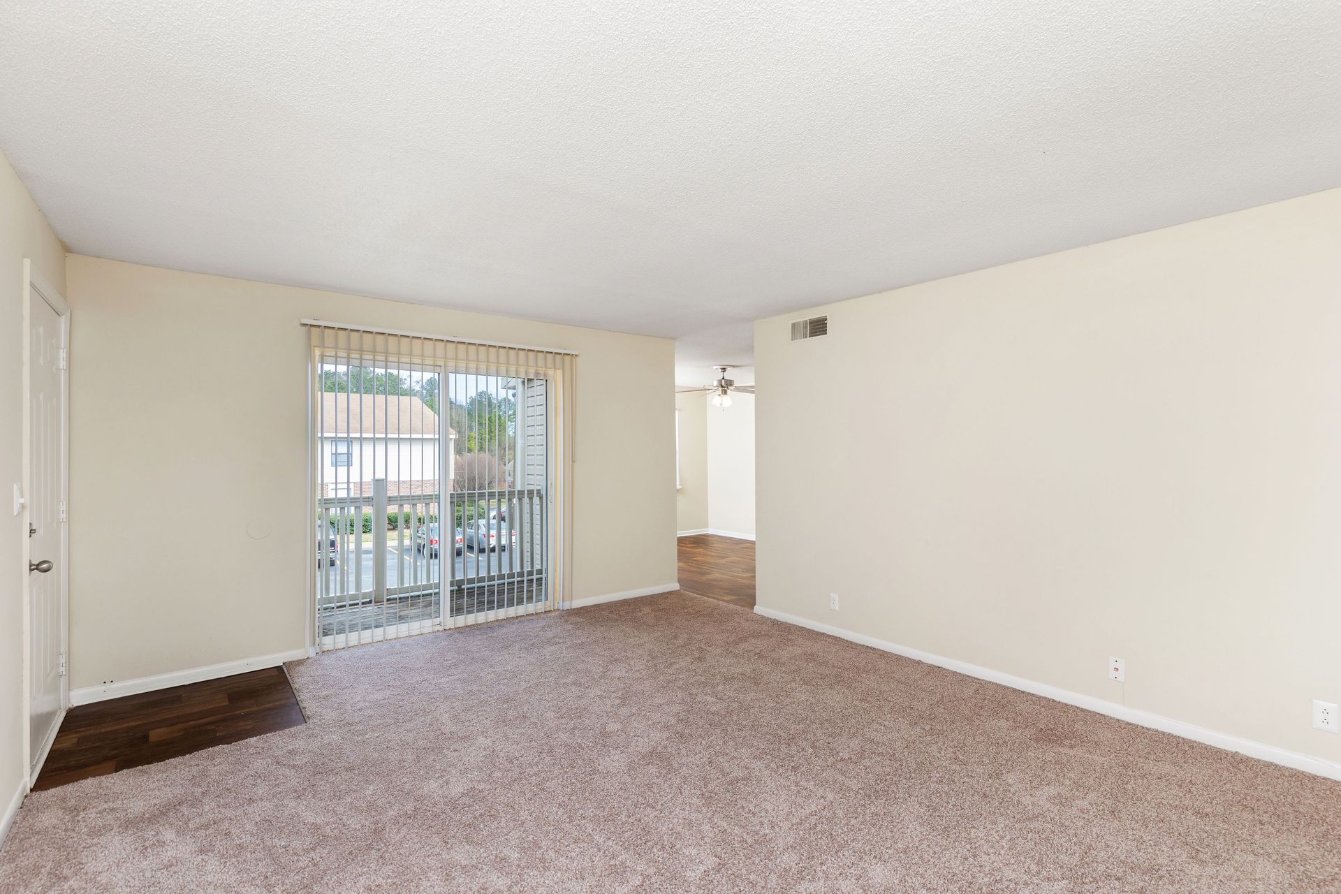 Empty living room with beige carpet, white walls, and a sliding glass door leading to a balcony.