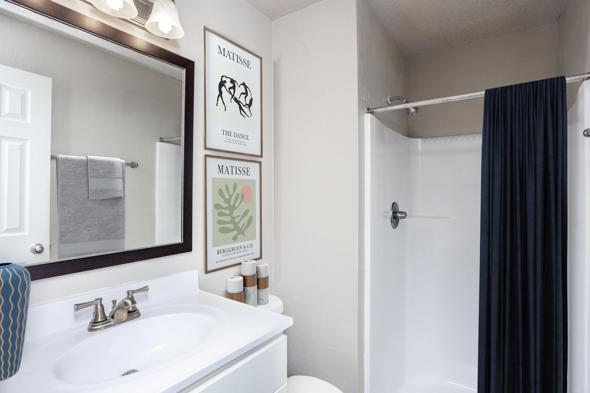A modern bathroom with a white vanity, a dark-framed mirror, two framed art prints, and a dark shower curtain.
