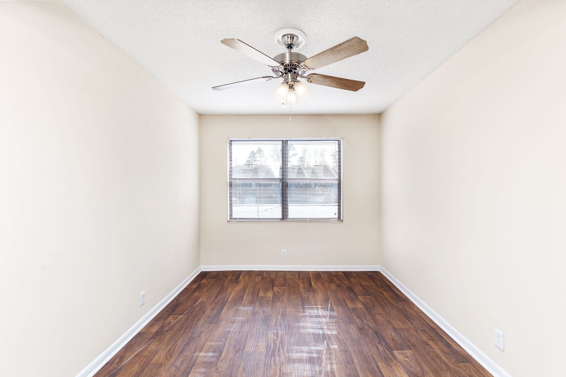 An empty room with plain beige walls, dark wood-look floors, a ceiling fan, and a single window with blinds.
