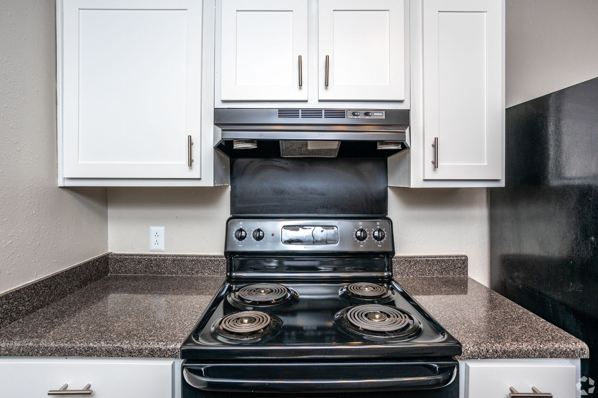 A kitchen stove with a range hood, brown speckled countertops, and white upper cabinets.