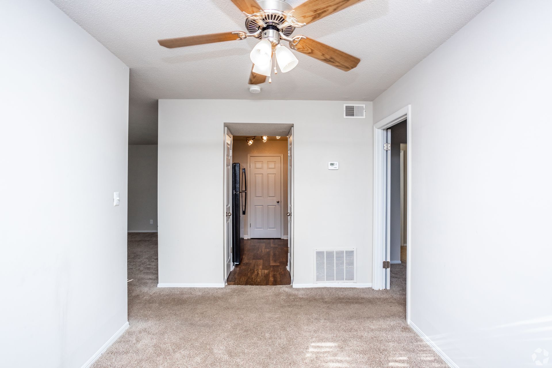A neutral-toned room with beige carpet, a ceiling fan, and doorways leading to a kitchen and other rooms.