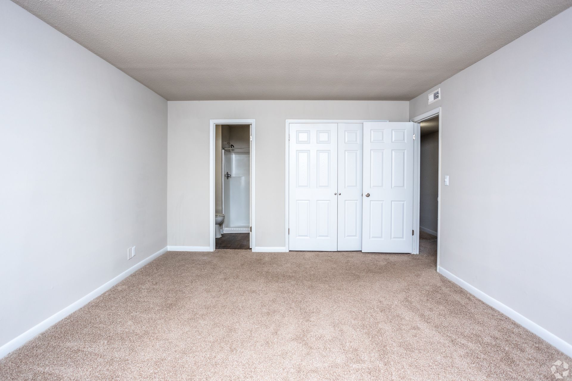 A bedroom with beige carpet, light gray walls, a popcorn ceiling, a bathroom doorway, and white double closet doors.