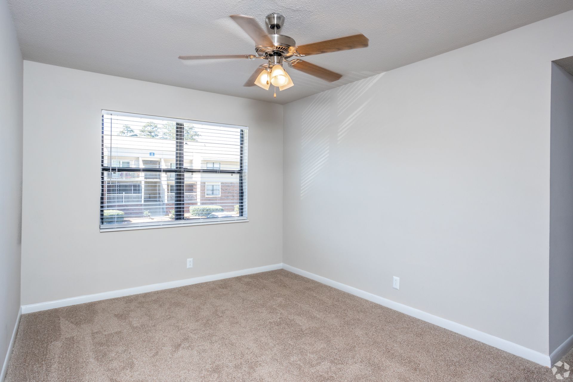 An empty bedroom with beige carpet, light-colored walls, a ceiling fan, and a window with blinds.