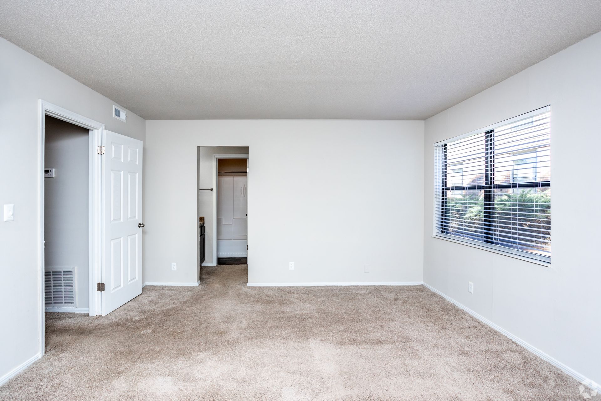 Empty room with beige carpet, white walls, a doorway to another room, a closet door, and a window with blinds.