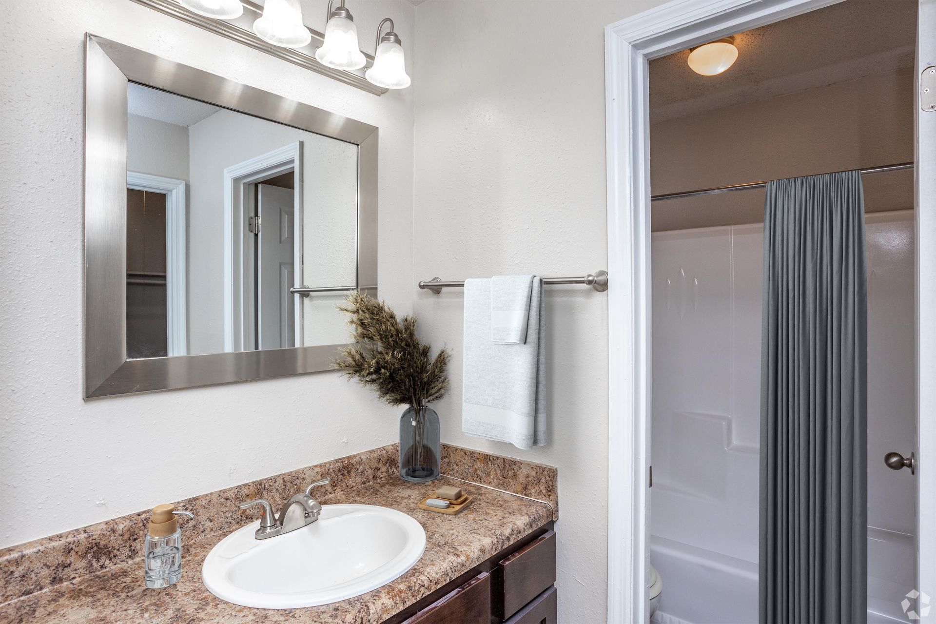 A bathroom vanity with a speckled countertop, white sink, framed mirror, wall light, and a shower with a gray curtain.