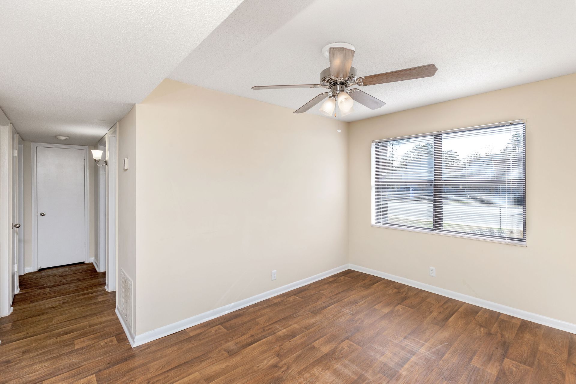 An empty, brightly lit room with beige walls, wood-look flooring, a ceiling fan, and a window with horizontal blinds.