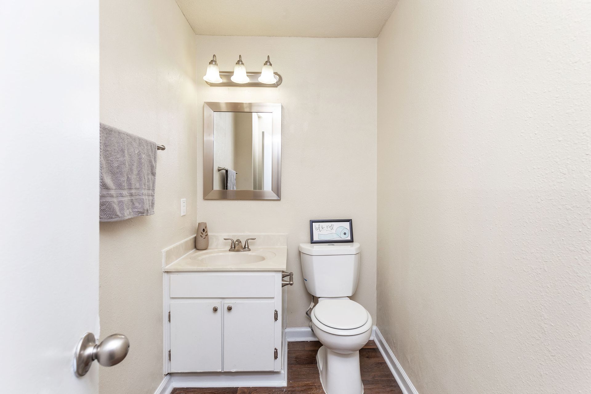 A small, neutral-toned bathroom featuring a white vanity cabinet, a wall-mounted mirror with lights, and a white toilet.