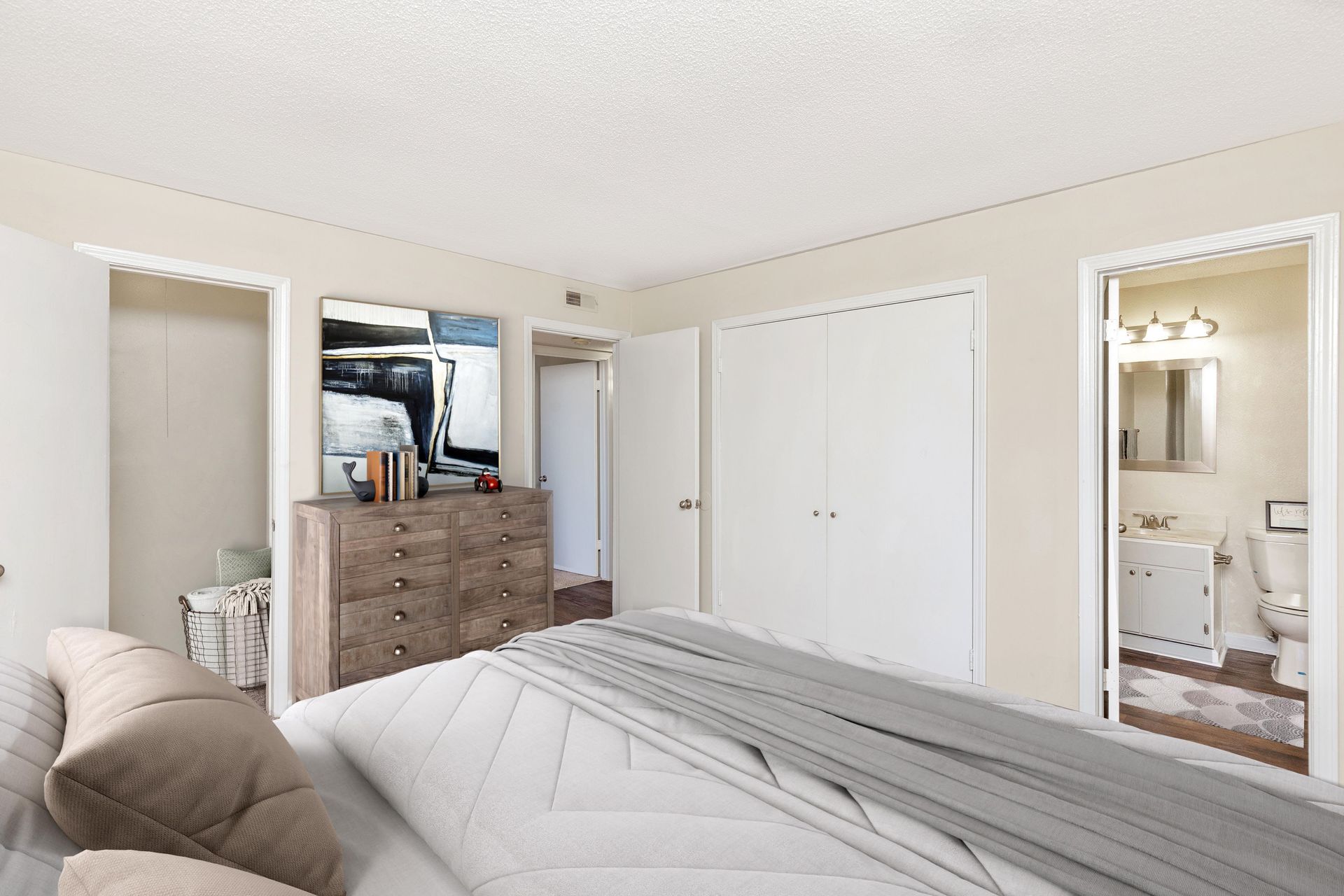 A bedroom featuring a bed with gray bedding, a wood dresser with abstract art, white closet doors, and an en suite bath.